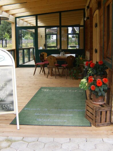 An inviting entrance area of a café with wooden decor. Flowers in pots enhance the atmosphere, and tables are visible inside. | © Speckhütte