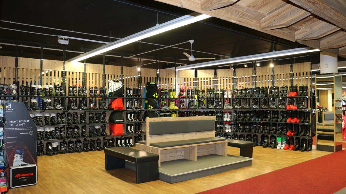 Ein modernes Sportgeschäft mit zahlreichen Skischuhen und Ausrüstungen an den Wänden. In der Mitte befindet sich eine Sitzgelegenheit aus Holz. | © Sport 2000 Rankl Schladming