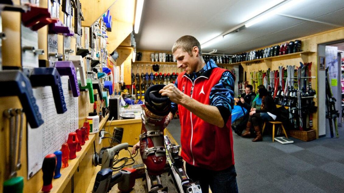Ein Mitarbeiter in einem Skishop repariert eine Skischuhe. Im Hintergrund sind verschiedene Skiausrüstungen und eine gemütliche Wartezone zu sehen. | © Sport Schöttl