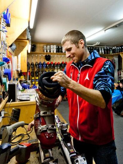 An employee in a ski shop is repairing a ski boot. In the background, various ski equipment and a cozy waiting area can be seen. | © Sport Schöttl