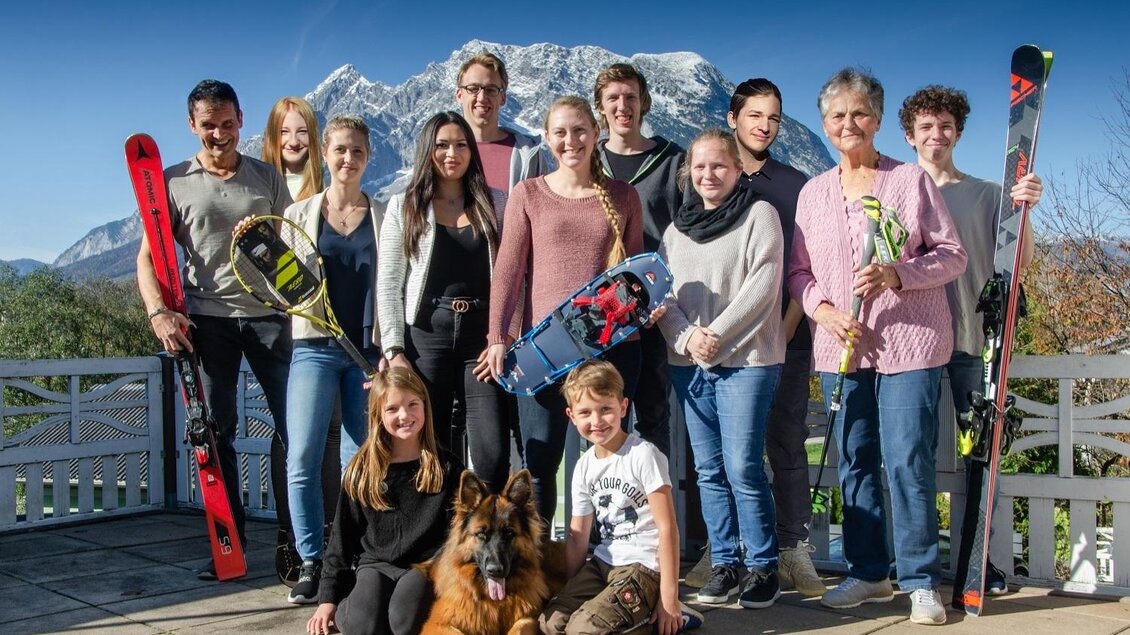 Eine Gruppe von Menschen steht auf einer Terrasse mit Blick auf die Berge. Einige halten Skier, während ein Hund davor sitzt. | © Sport Schöttl
