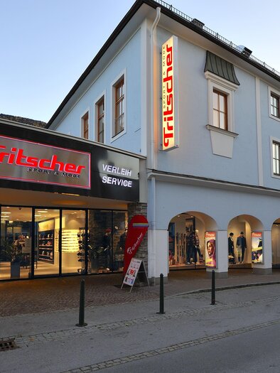 Das Bild zeigt das Sportgeschäft Tritscher mit beleuchtetem Eingangsbereich und Wintersport-Schaufenstern am frühen Abend. | © Sport Tritscher