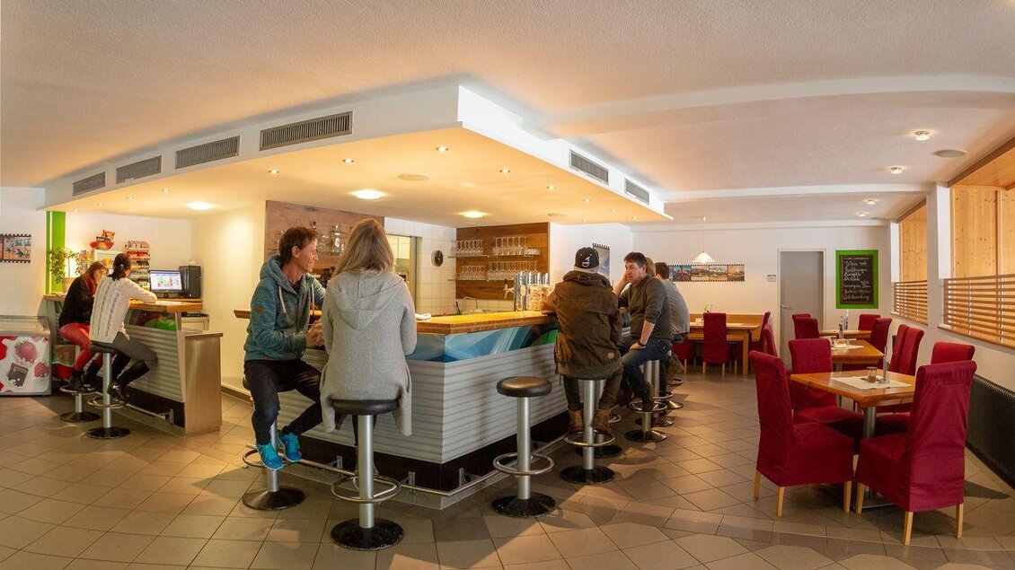 Eine gemütliche Bar mit Sitzplätzen an der Theke und roten Sesseln. Im Hintergrund sind Gäste beim Entspannen und Unterhalten zu sehen. | © Sportsarea Grimming