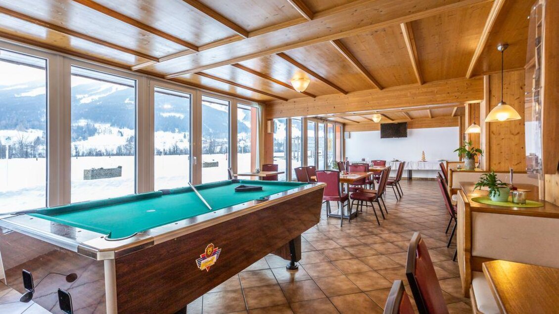 Ein gemütlicher Raum mit einem Billardtisch und großen Fenstern, die einen Blick auf die schneebedeckten Berge bieten. Es gibt Tische und Stühle für Gäste, die zum Entspannen einladen. | © Sportsarea Grimming
