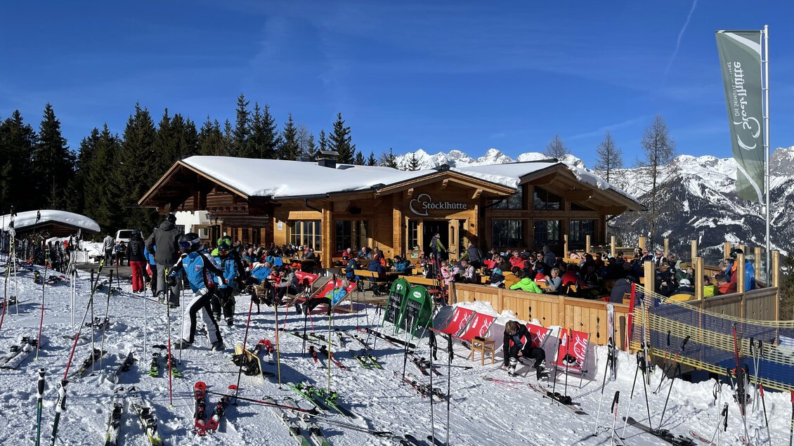 Eine Skihütte umgeben von frisch gefallenem Schnee. Viele Menschen entspannen sich auf der Terrasse und genießen die Sonne. | © Stöcklhütte