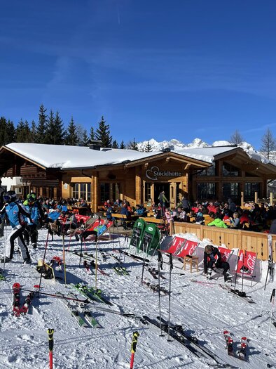 A ski cabin surrounded by freshly fallen snow. Many people are relaxing on the terrace and enjoying the sun. | © Stöcklhütte