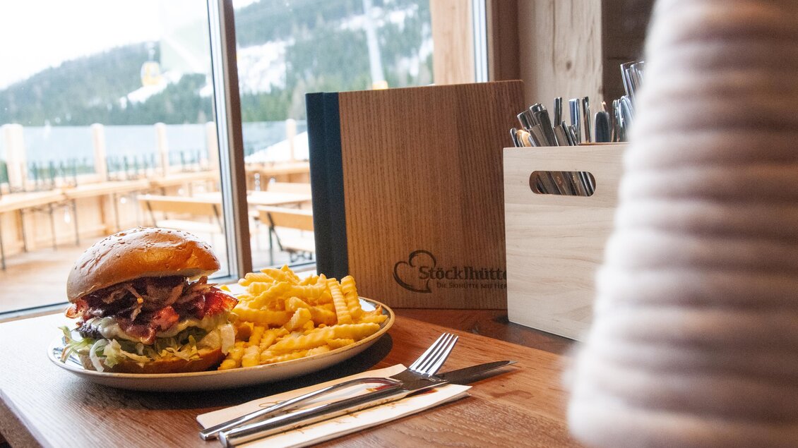 Ein leckerer Burger mit frischem Gemüse und knusprigen Pommes liegt auf einem Tisch. Im Hintergrund sind Fenster mit Bergblick zu sehen. | © Stöcklhütte