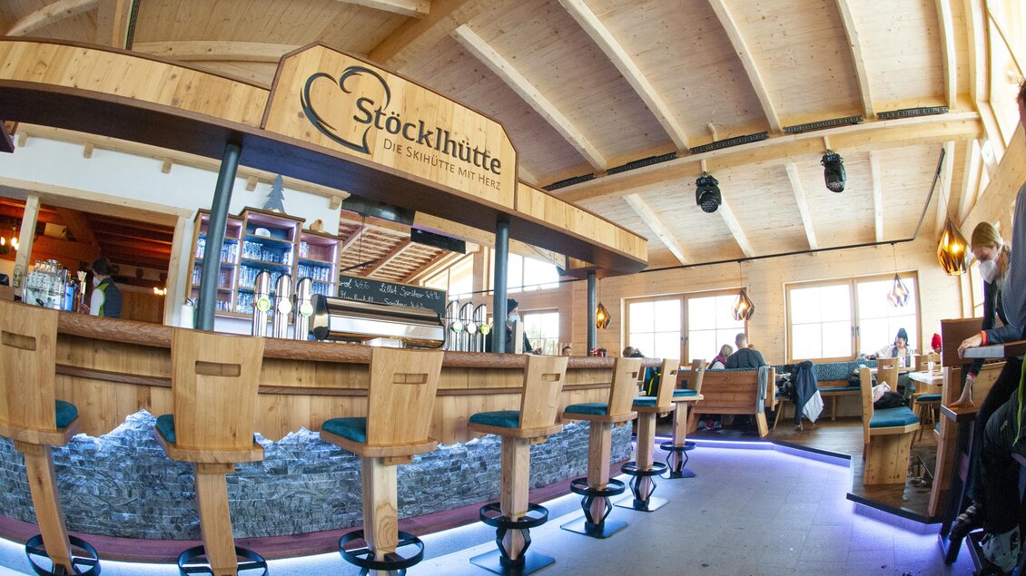 Eine gemütliche Bar mit Holzverkleidung und Beleuchtung. Im Hintergrund sind Bänke und Fenster, die für eine einladende Atmosphäre sorgen. | © Stöcklhütte