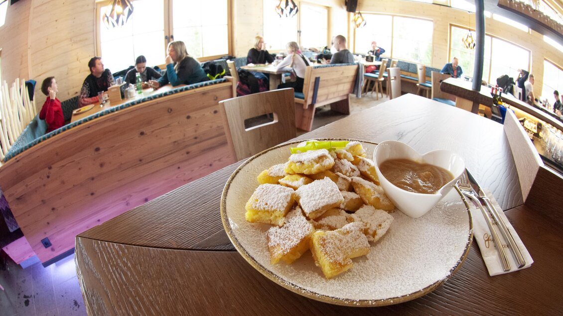 Ein Teller mit gebackenem Gebäck und Soße steht auf einem Holztisch. Im Hintergrund sieht man Menschen, die in einem gemütlichen Restaurant sitzen. | © Stöcklhütte