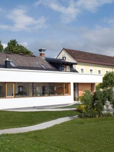Ein modernes Haus mit großen Fenstern in einem grünen Garten. Im Hintergrund sind Berge und weitere Gebäude sichtbar. | © Stüberl - Cafè am Kurpark