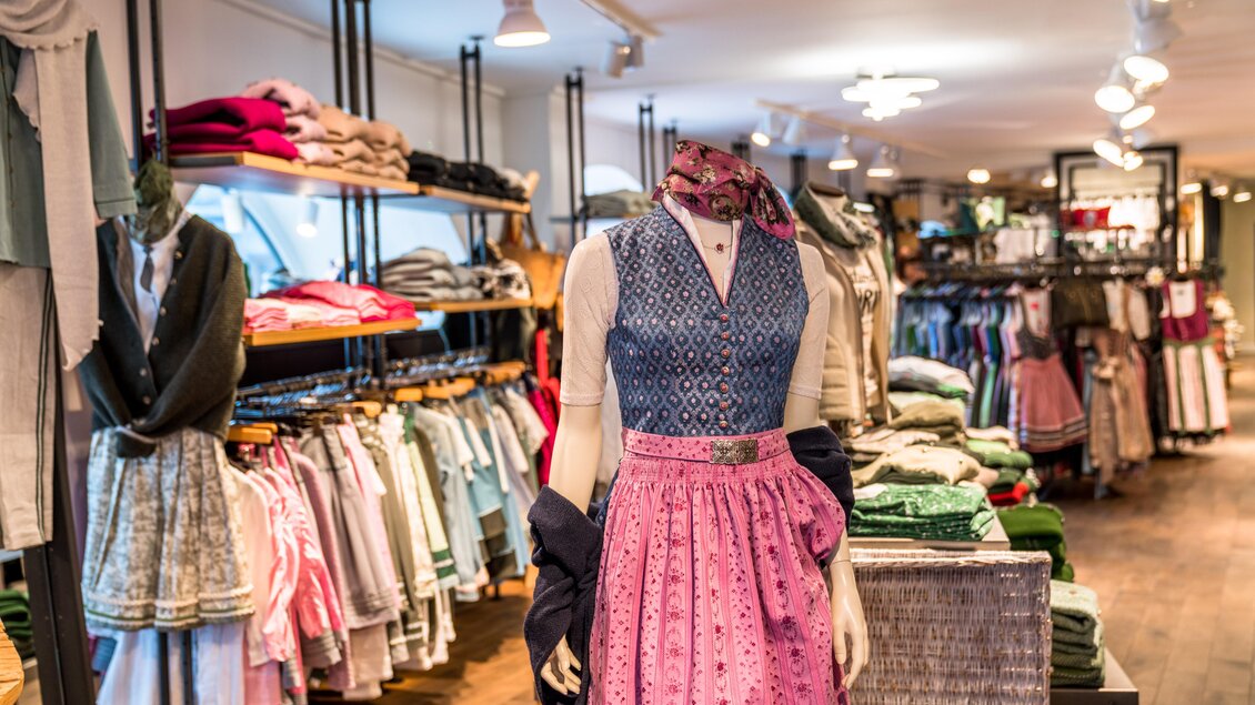 Ein Geschäft mit traditioneller Trachtenmode. Im Vordergrund steht eine Schneiderpuppe in einem bunten Dirndl, umgeben von vielen Kleidungsstücken. | © Steirerland Tracht & Leder