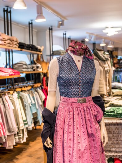 A shop with traditional folk costumes. In the foreground stands a dressmaker's dummy in a colorful dirndl, surrounded by many garments. | © Steirerland Tracht & Leder