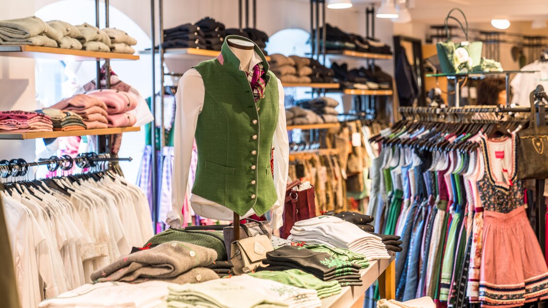 Ein bekleideter Mannequin in einer Tracht aus grünem Stoff steht im Vordergrund. Im Hintergrund sind Regale mit verschiedenen Kleidungsstücken und Accessoires zu sehen. | © Steirerland Tracht & Leder
