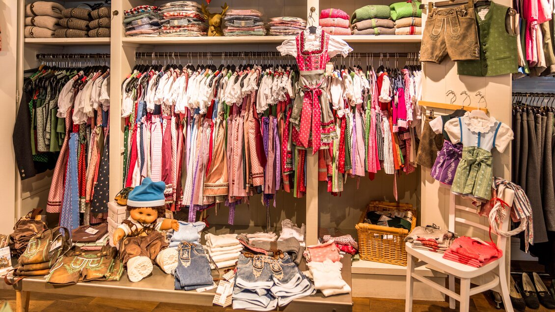 Ein bunter Bekleidungsladen mit einer Vielzahl von Kinderbekleidung hängt an einer Wand. Auf einem Tisch sind weitere Kleidungstücke und einige Spielzeuge ausgelegt. | © Steirerland Tracht & Leder