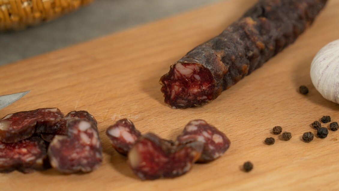 Eine Wurst auf einem Holzbrett, teilweise angeschnitten. Neben der Wurst liegen einige Scheiben und Gewürzperlen. | © Perner Alexandra