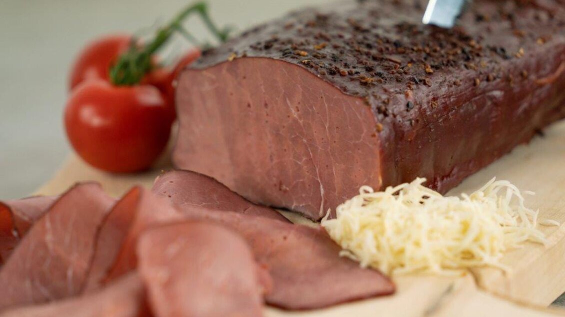 Ein Stück saftiges Fleisch auf einem Holzbrett, neben frisch geschnittenen Scheiben. Im Hintergrund sind einige Tomaten und Kräuter zu sehen. | © Perner Alexandra