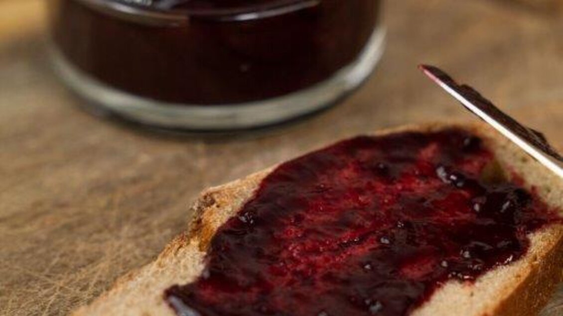 Ein Stück Brot mit dunkelroter Marmelade und ein Glas mit Marmelade im Hintergrund. Die Marmelade ist gleichmäßig auf dem Brot verteilt. | © Perner Alexandra