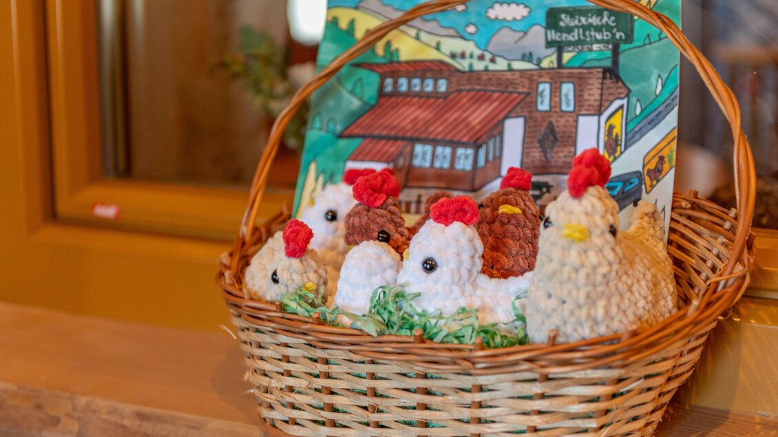 Ein Korb mit liebevoll gehäkelten Hühnern in verschiedenen Farben. Im Hintergrund ist eine bunte Zeichnung eines Bauernhauses zu sehen. | © Steirische Hendlstub'n