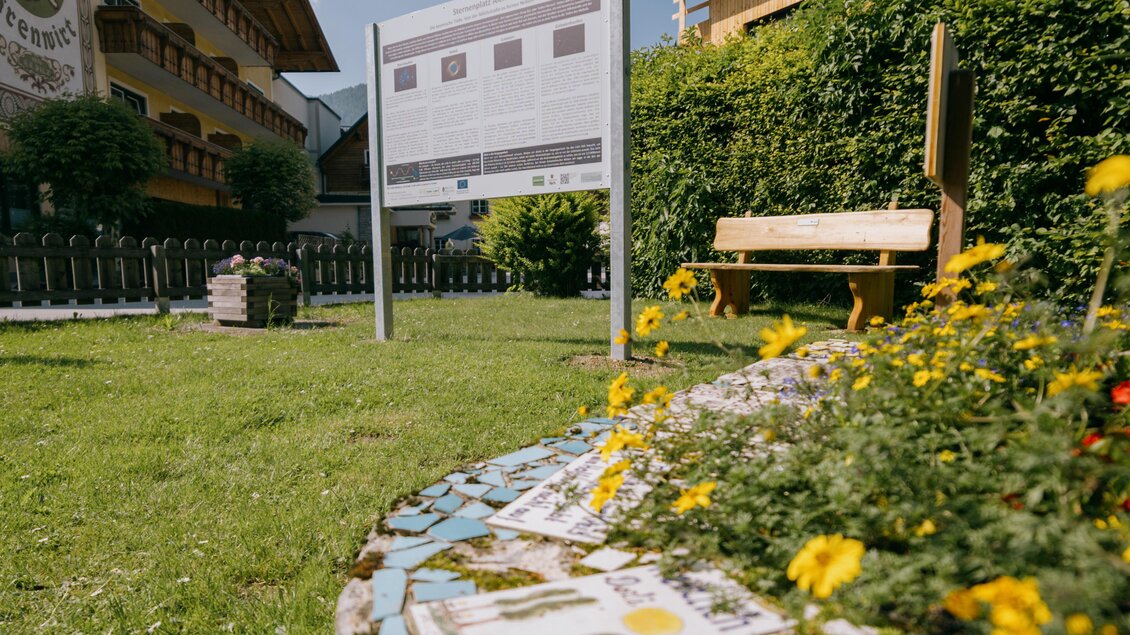 Eine grüne Wiese mit bunten Blumen und einem Mosaikrand. Im Hintergrund steht ein Informationsschild und ein Holzbank. | © Gerald Grünwald