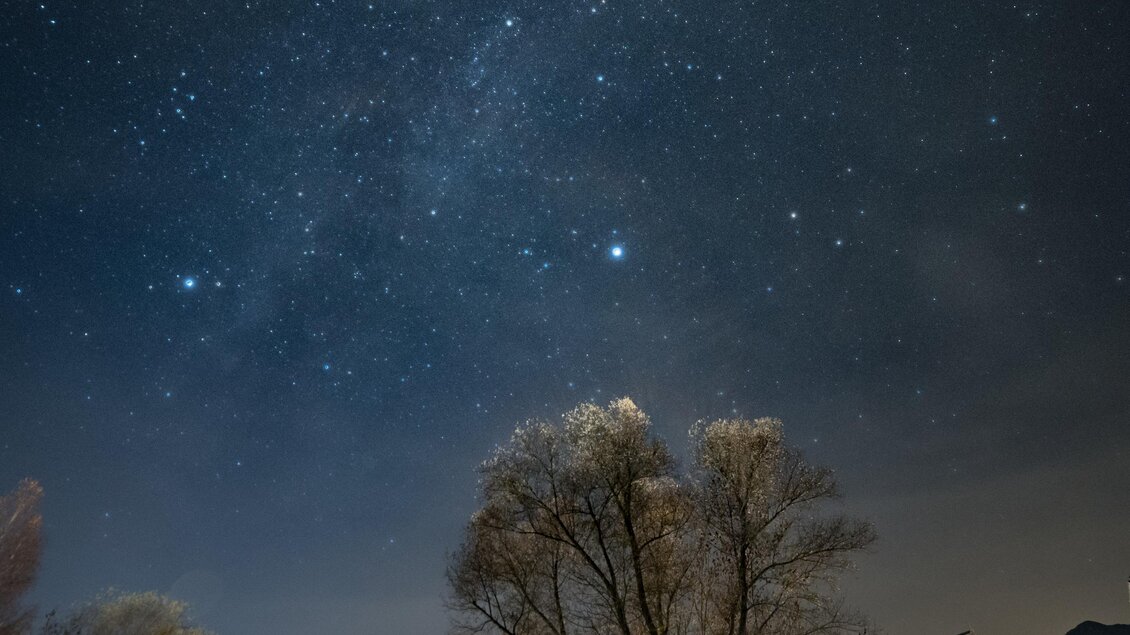 Ein malerischer Nachthimmel mit vielen Sterne und einer schönen Unterkunft im Vordergrund. Unten sind Bäume und ein ruhiges Gewässer zu sehen. | © Gerald Grünwald