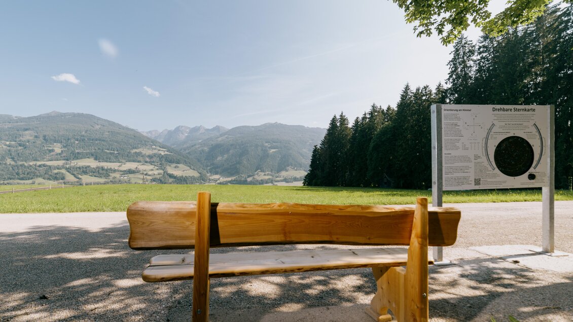 Eine ruhige Bank steht vor einer beeindruckenden Berglandschaft. Im Hintergrund sind Wiesen und Wälder zu sehen, während ein Informationsschild in der Nähe steht. | © Gerald Grünwald