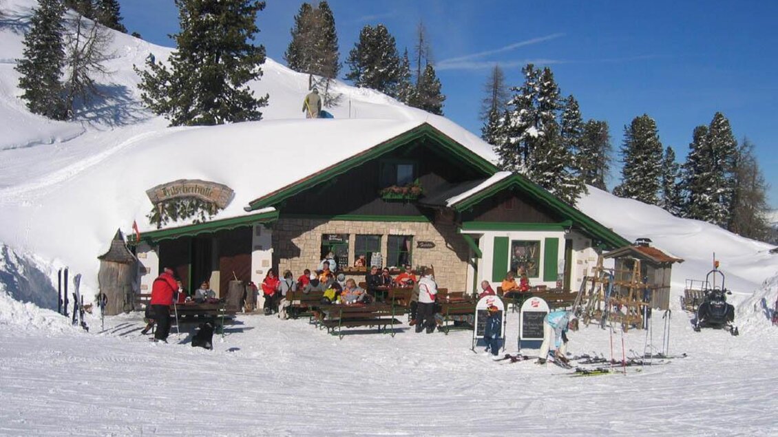 Eine gemütliche Berghütte im Schnee mit Gästen auf der Terrasse. Im Hintergrund sind schneebedeckte Bäume und ein klarer blauer Himmel zu sehen. | © Stoderhütte
