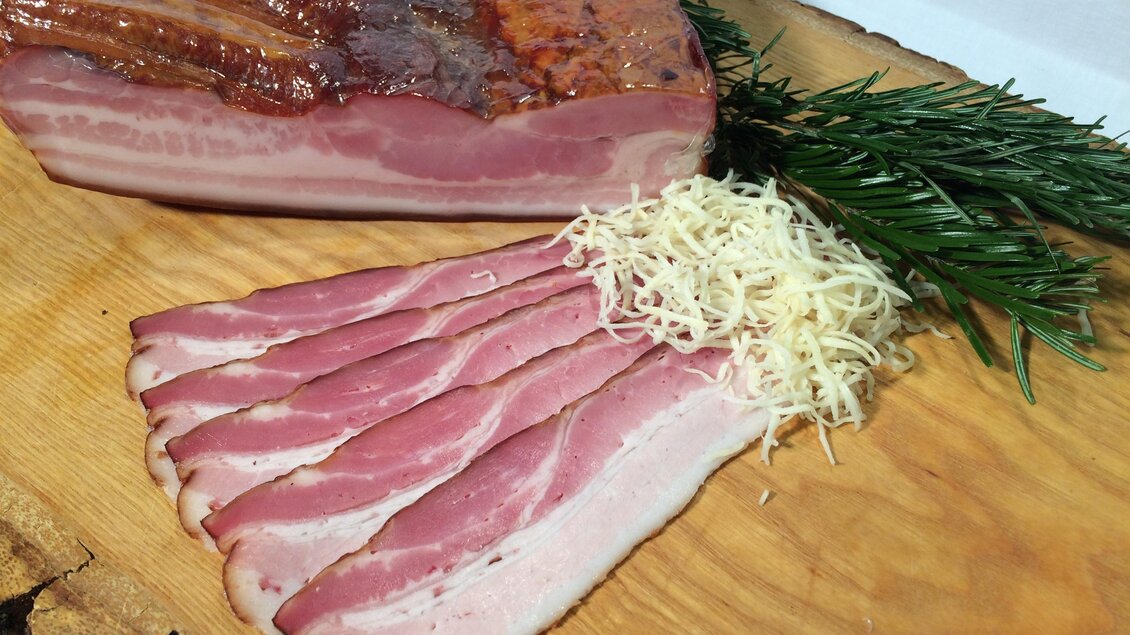 Ein Stück geräucherter Speck liegt auf einem Holzbrett. Neben dem Speck sind einige dünne Scheiben und frische Kräuter platziert. | © Andreas Spanberger