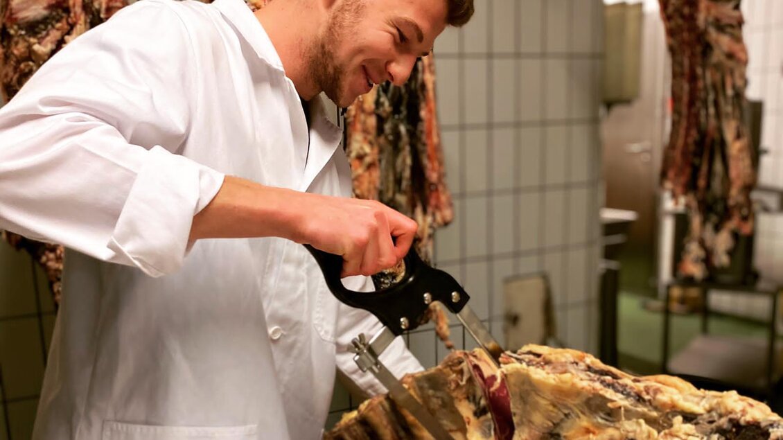 Ein Metzger schneidet Fleisch mit einer Säge in einer Fleischerei. Im Hintergrund hängen verschiedene Fleischstücke. | © Andreas Spanberger
