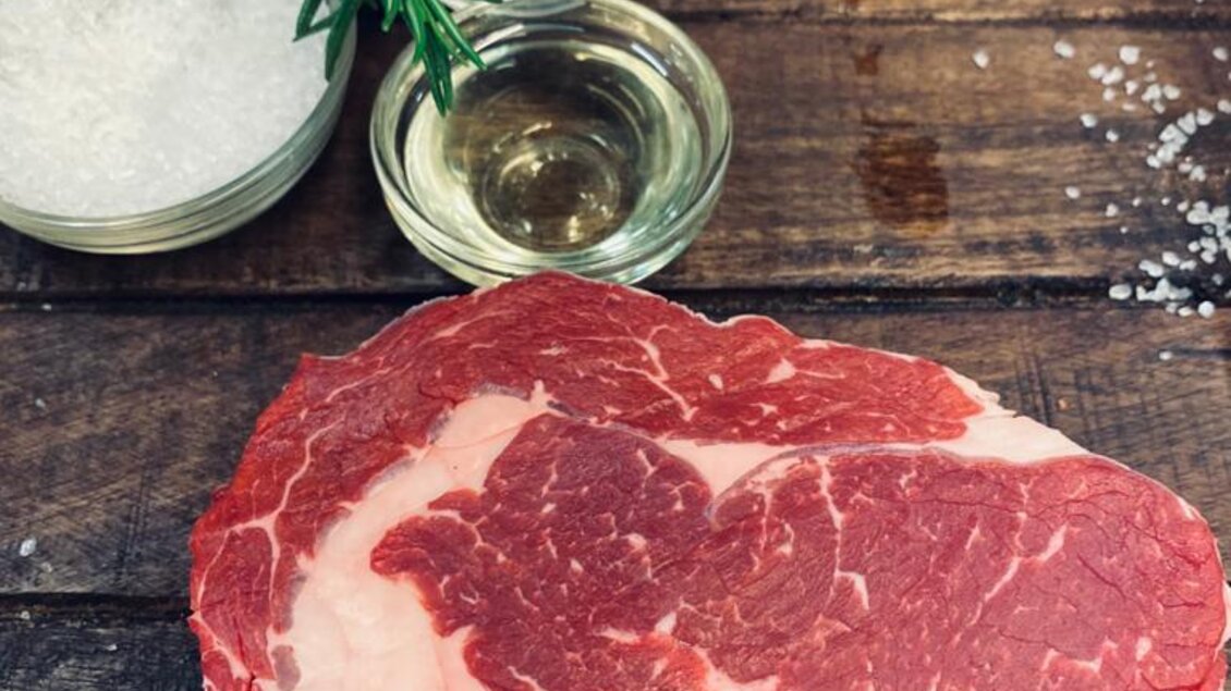 Ein frisches Stück Fleisch auf einem Holztisch, umgeben von Salz, Pfefferkörnern und Kräutern. Im Hintergrund ist das Logo einer Fleischerei zu sehen. | © Andreas Spanberger