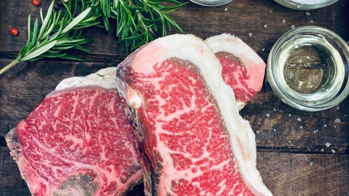 Zwei saftige Fleischstücke mit feiner Marmorierung liegen auf einem Holzbrett. Daneben sind Kräuter, Gewürze und Öl dekorativ angeordnet. | © Andreas Spanberger
