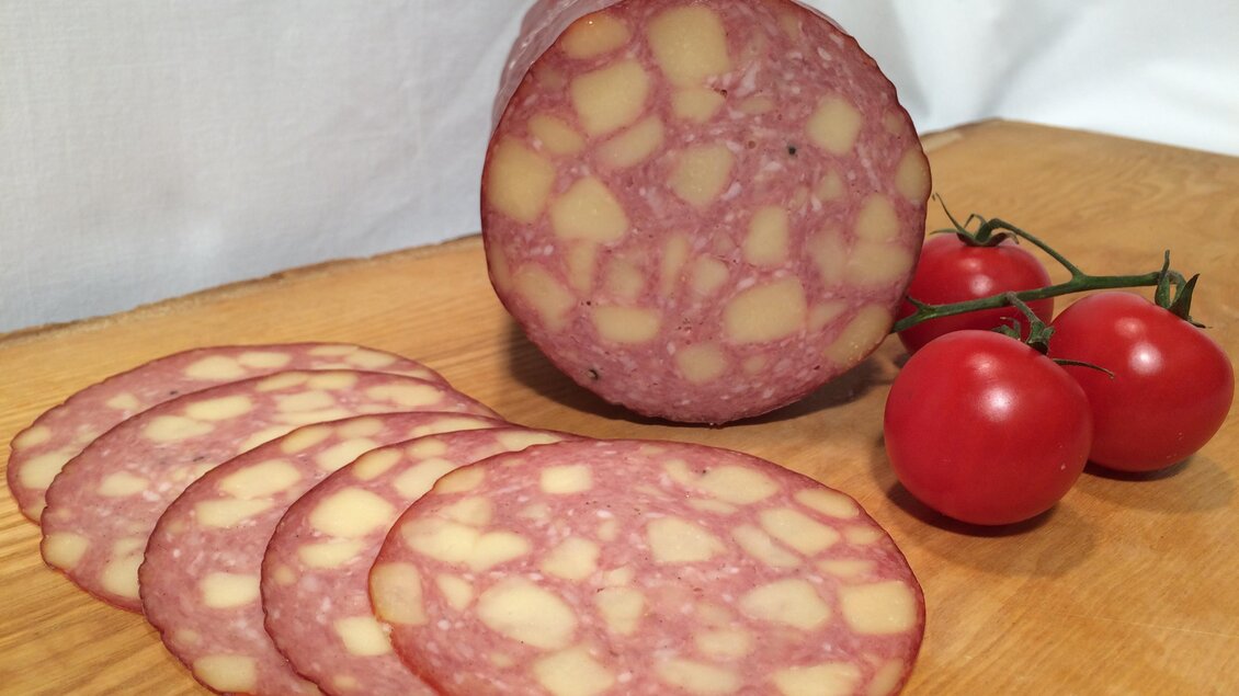 Eine Scheibe Wurst mit sichtbaren Fettstückchen liegt auf einem Holzbrett. Daneben befinden sich einige frische, rote Tomaten. | © Andreas Spanberger