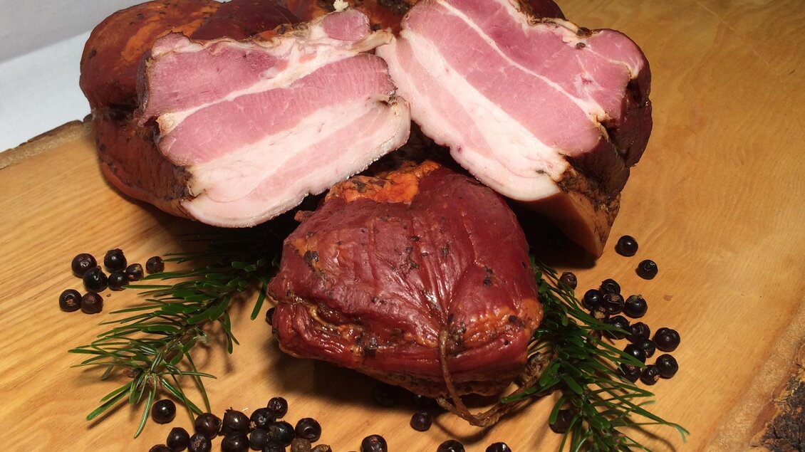 Eine Auswahl von geräuchertem Fleisch auf einem Holzbrett. Daneben liegen hartgekochte schwarze Beeren und etwas frisches Grün. | © Andreas Spanberger