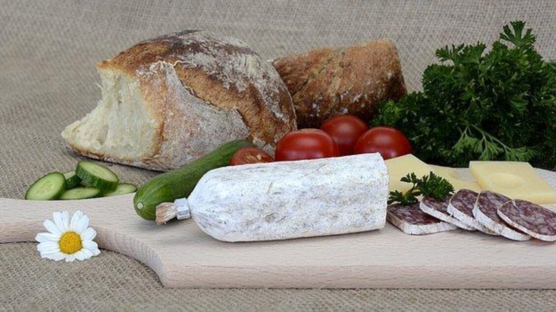 Eine Holzplatte mit verschiedenen Lebensmitteln: frisches Brot, Wurst, Käse, Tomaten, Gurken und etwas Petersilie. Alles ist ansprechend angeordnet und vermittelt ein gemütliches Picknickgefühl. | © Ochsenbeisl Spanberger