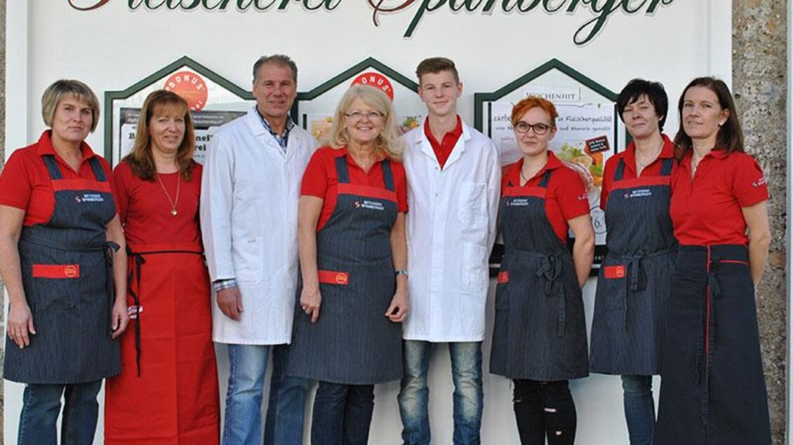Eine Gruppe von Mitarbeitern der Fleischerei Spanberger steht vor ihrem Geschäft. Sie tragen einheitliche Outfits und zeigen ein freundliches Lächeln. | © Ochsenbeisl Spanberger