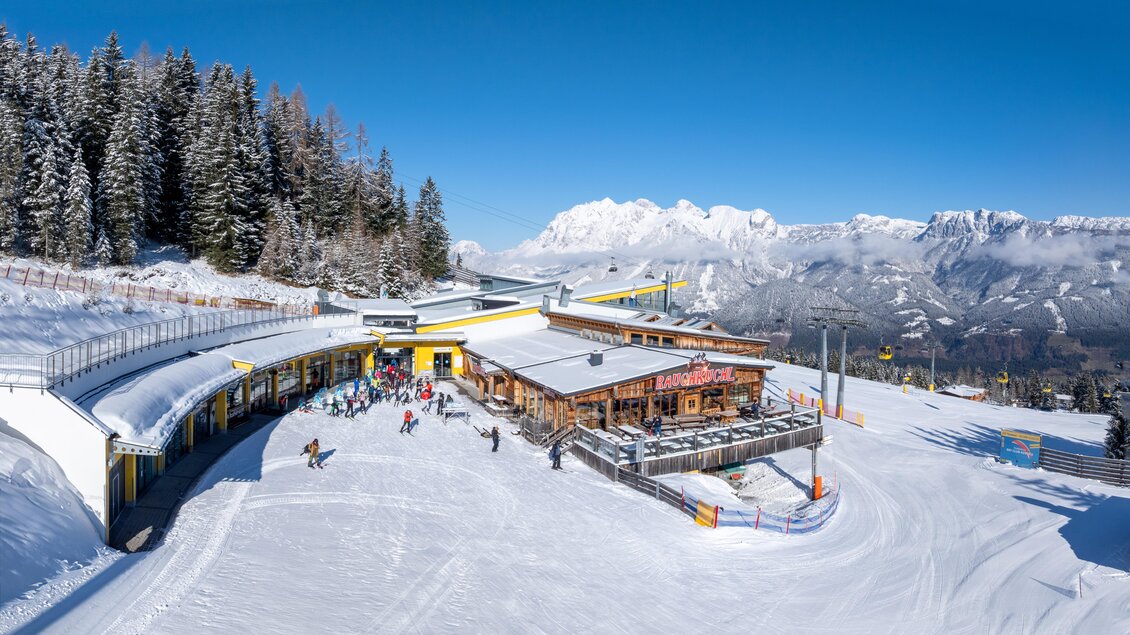 Skifahrer genießen die Sonnenterrasse der Stoni´s Rauchkuchl mit Blick auf die verschneiten Berge. Daneben die Gondelbahn und weitläufige Skipisten. | © Stoni´s Rauchkuchl