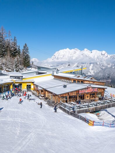 Skiers enjoy the sun terrace of Stoni's Rauchkuchl with a view of the snow-covered mountains. Next to it, the gondola lift and extensive ski slopes. | © Stoni´s Rauchkuchl