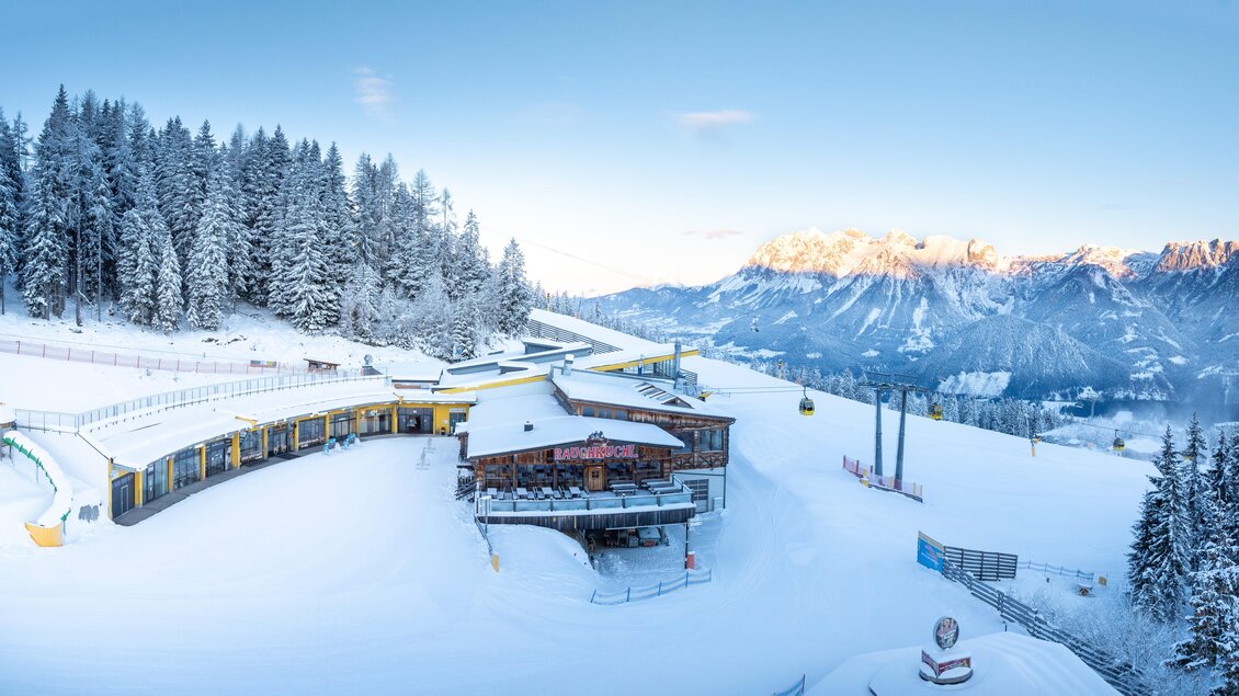 Panoramablick auf die breiten Skipisten am Hauser Kaibling. Unten im Bild liegt die Rauchkuchl neben der Gondelbahn, umgeben von verschneiten Wäldern. | © Stoni´s Rauchkuchl