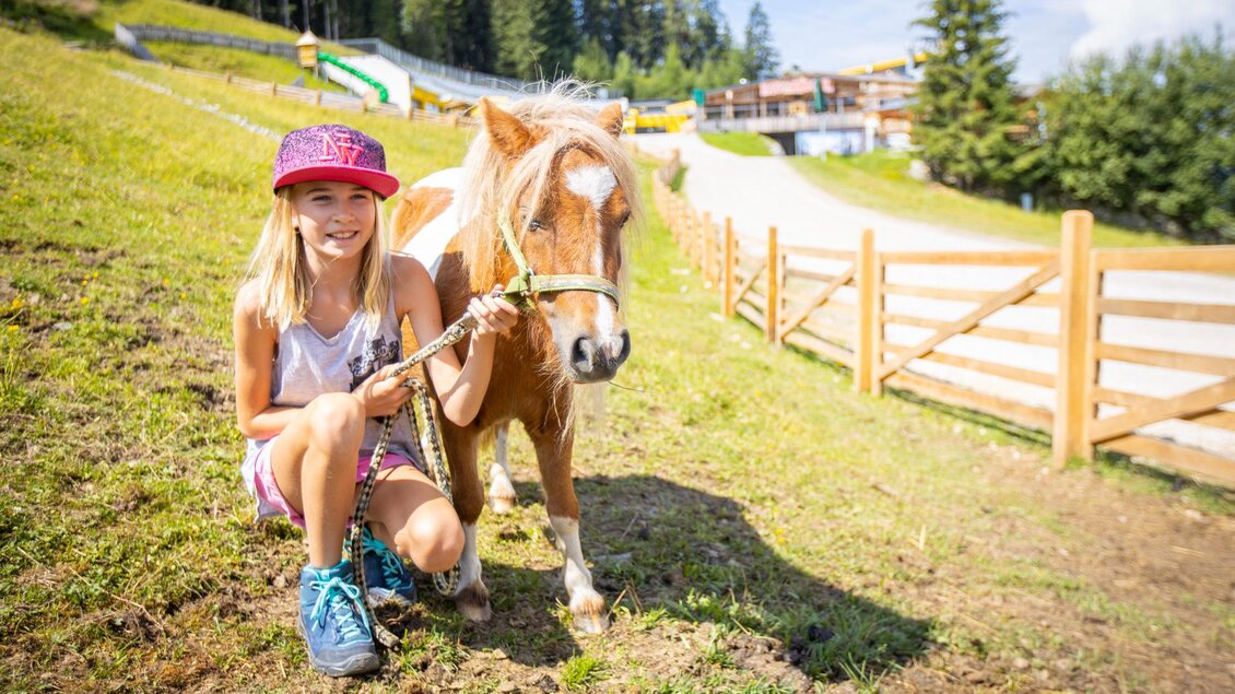 Ein Mädchen mit einer pinken Kappe sitzt neben einem kleinen Pony auf einer Wiese. Im Hintergrund sind Bäume und eine Holzumzäunung zu sehen. | © Haus Kaibling