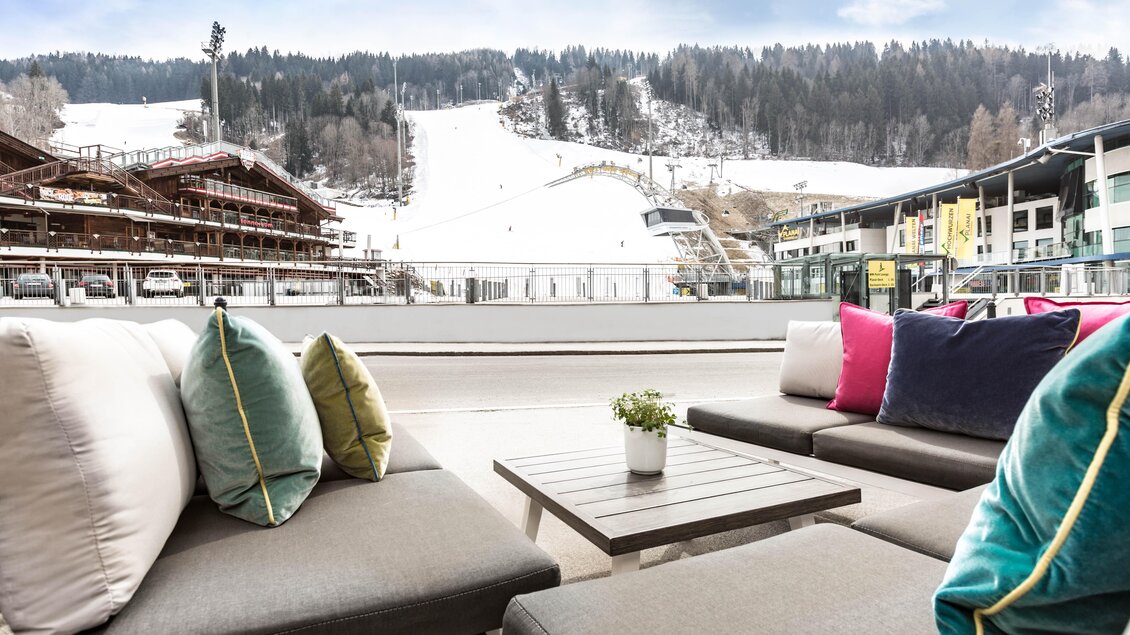 Eine gemütliche Lounge mit modernen Sofas und bunten Kissen. Im Hintergrund sieht man eine schneebedeckte Piste und Wälder. | © TUI BLUE