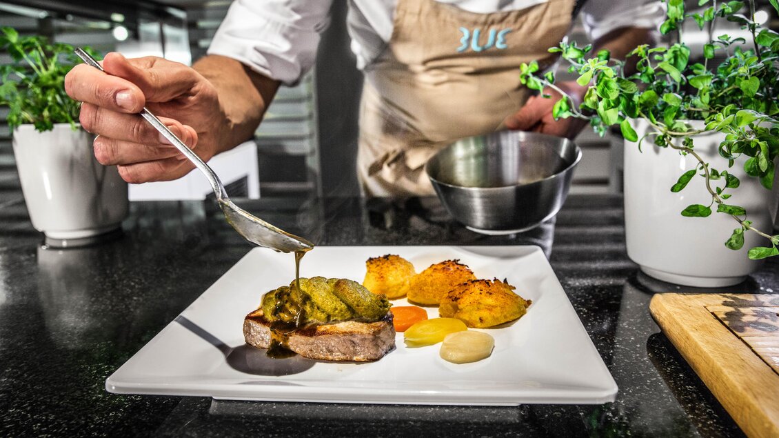 Ein Koch bereitet ein kunstvoll angerichtetes Gericht auf einem weißen Teller vor. Frisches Gemüse und Soße werden hinzugefügt, um das Gericht zu vervollständigen. | © TUI BLUE