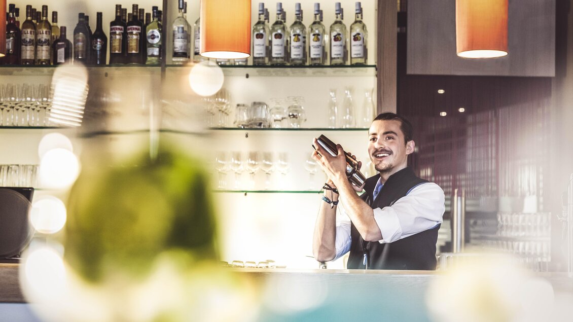 Ein Barkeeper lächelt und schüttelt einen Cocktailshaker hinter der Bar. Im Hintergrund sind Spirituosenflaschen gut sichtbar. | © TUI BLUE