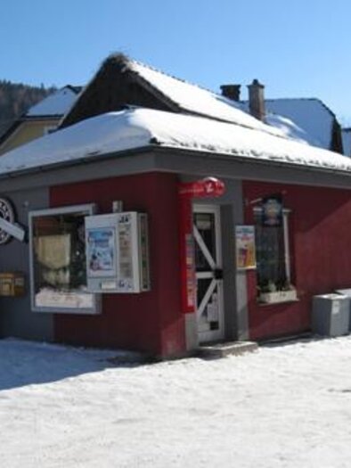 A small red shop with a snow-covered roof and various offerings on the facade. Surrounded by snow and a clear blue sky.