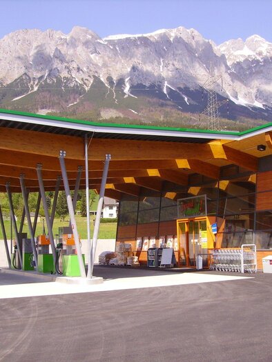 Ein modernes Gebäude mit Holzdetails steht vor einer beeindruckenden Bergkulisse. Der Himmel ist klar und die Umgebung ist grün. | © Tankstelle - Lebensmittelgeschäft - Bistro - Land lebt auf!