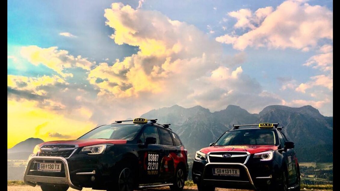 Zwei Taxis stehen auf einer Straße vor einer malerischen Berglandschaft. Der Himmel ist teilweise bewölkt, mit beeindruckenden Farben bei Sonnenuntergang. | © Taxi Bernd