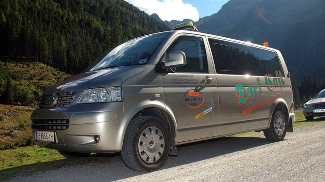 Ein silberner Transporter parkt auf einer malerischen Straße umgeben von Bergen und Wald. Der Himmel ist klar und die Umgebung ist grün und einladend. | © Taxi Heli