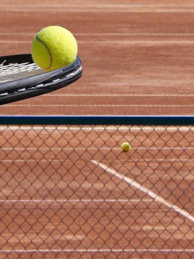 A tennis ball hovers over a tennis racket on a sand court. In the background, another tennis ball can be seen. | © Jagdhof