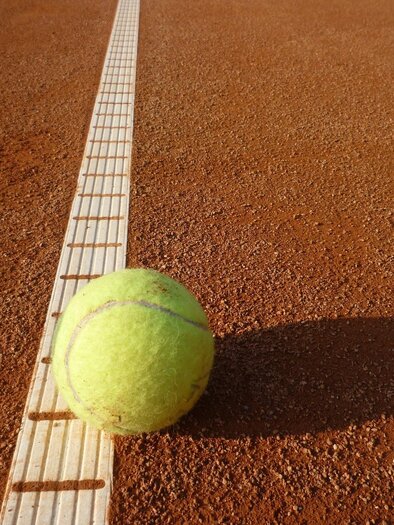 A tennis ball lies on a clay court next to the line. The ground is red and the sun is shining. | © Hans Dietmann (Pixabay)