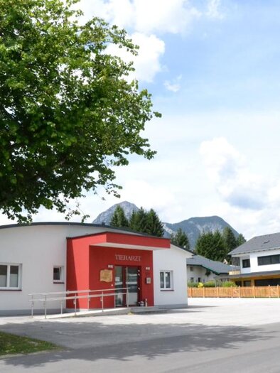 A modern building with a red facade stands by a road. In the background, green trees and mountains can be seen. | © Dr. Robert Gruber