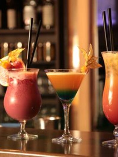 Drei bunte Cocktails stehen auf einer Bar. Der Hintergrund zeigt mehrere Flaschen und schafft eine gemütliche Atmosphäre. | © Fotolia