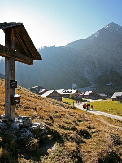 Eine malerische Berglandschaft mit einem Holzschild im Vordergrund. Im Hintergrund sind Hütten und Wanderer auf einem schmalen Weg zu sehen. | © Herbert Raffalt
